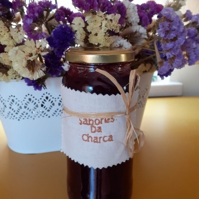 Garrafa de vidro com geleia vermelha e etiqueta de tecido com texto 'Sabores Da Charca' ao fundo flores secas em vaso branco.