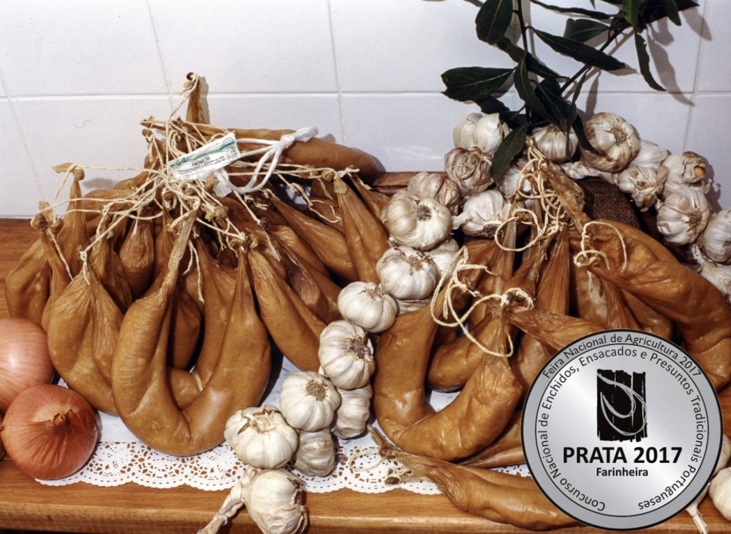Farinheira da Beira-Baixa (2 un.) Chouriços curados, cachos de alho e cebolas numa mesa com toalha rendada e azulejos brancos ao fundo.