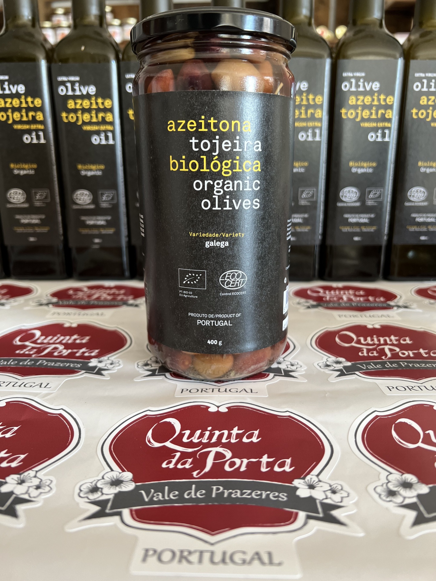 Azeitona Galega Tojeira (BIO) 400g Frasco de azeitonas biológicas Tojeira com rótulo preto sobre superfície com padrão Quinta da Porta