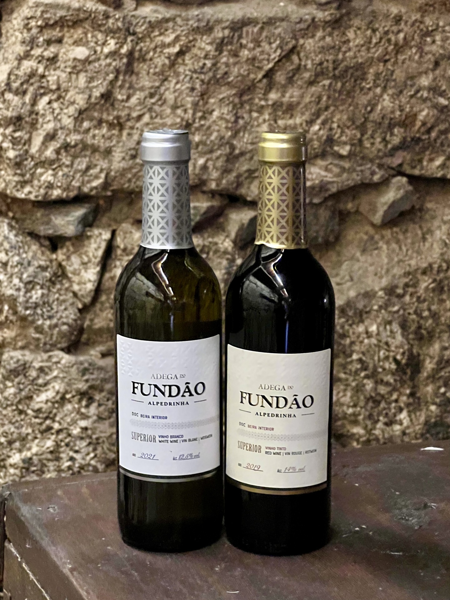 Alpedrinha (375ml) Duas garrafas de vinho ADEGA DO FUNDÃO ALPEDRINHA, uma branca e uma tinta, em superfície metálica com fundo de parede de pedra