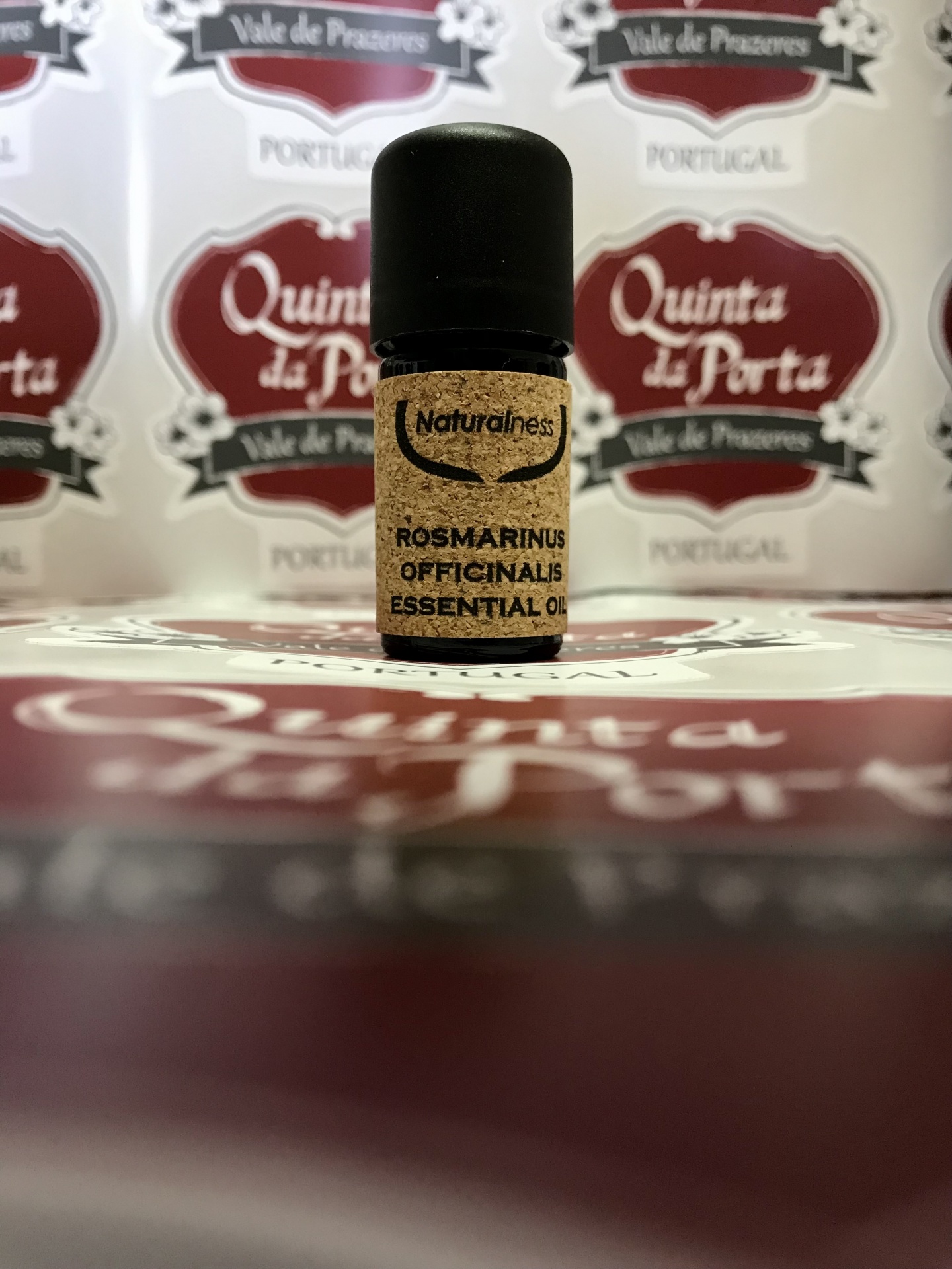 Óleo essencial de Alecrim (5ml) Frasco de óleo essencial com rótulo de cortiça e tampa preta, com fundo de etiquetas vermelhas e brancas.
