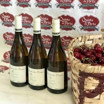 Três garrafas de vinho branco e um cesto com cerejas junto a um painel com logotipos da Quinta da Porta.