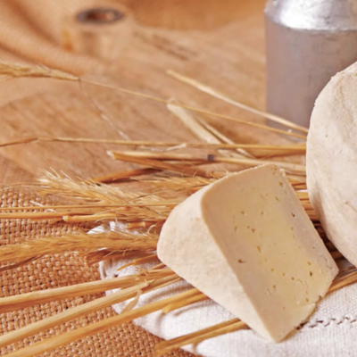 Pedaço de queijo amarelo sobre toalha branca com espigas de trigo e jarra metálica