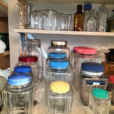 Frascos e jarros de vidro com tampas coloridas em estantes de madeira, ao fundo garrafas e copos de vidro