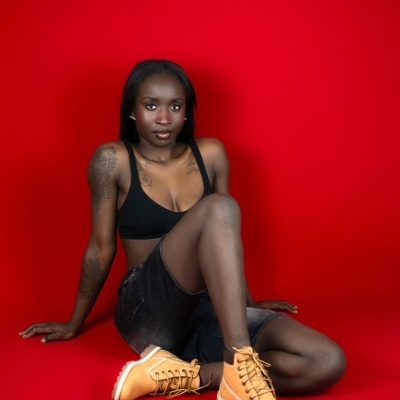 Mulher com top preto, calções de ganga pretos e botas amarelas sentada num fundo vermelho