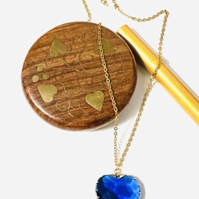 Colar dourado com pendente coração azul sobre caixa redonda de madeira com desenhos dourados