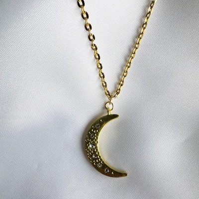 Colar dourado com pendente em forma de lua crescente com pedras brilhantes sobre tecido branco.