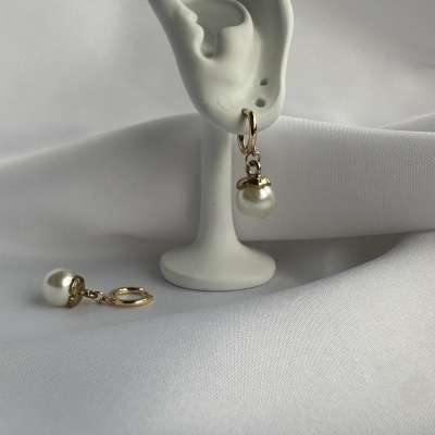 Par de brincos dourados com pérolas brancas em suporte de orelha branca sobre tecido branco.