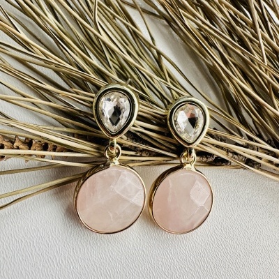 Brincos dourados com pedras transparente e rosa sobre superfície branca e ramos secos.