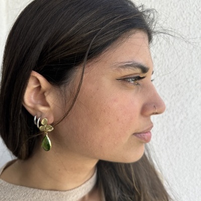 Pendentes dourados com pedra verde usados por mulher junto a parede branca