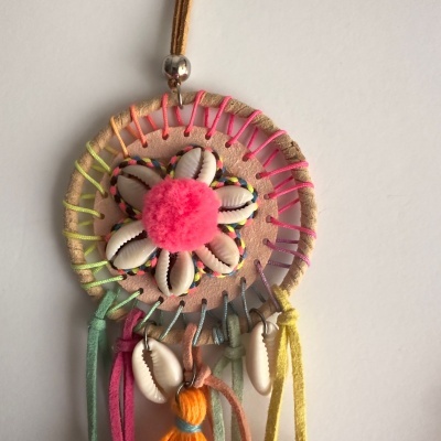 Pendente mandala com conchas, pompom rosa e fitas coloridas num fundo branco.