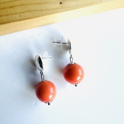 Brincos com esfera coral pendurados em base prateada sobre superfície branca e madeira clara