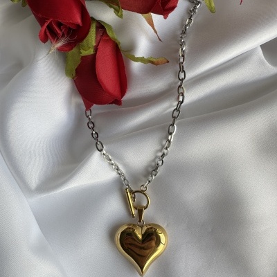 Colar prateado com pendente dourado em forma de coração, sobre tecido branco com rosas vermelhas