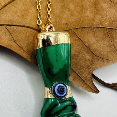Colar com pendente verde em forma de punho e detalhes dourados, com olho azul no centro, sobre uma folha seca