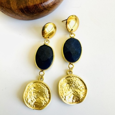 Brincos pendentes dourados com pedra preta e disco texturado em fundo branco próximo a suporte de madeira