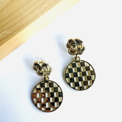 Brincos dourados com padrão xadrez pendentes sobre superfície branca e madeira