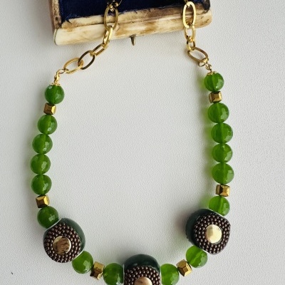 Colar com contas verdes e detalhes metálicos dourados sobre superfície branca junto a caixa azul de veludo
