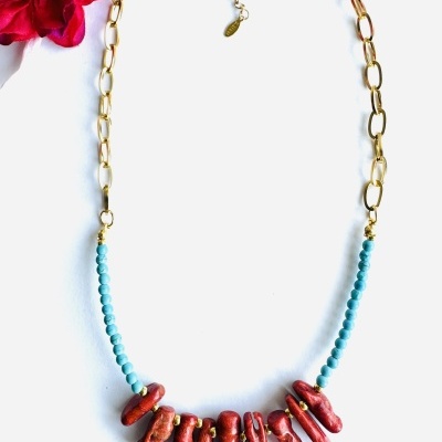 Colar dourado com contas azuis e pendentes vermelhos, sobre fundo branco com flor vermelha.