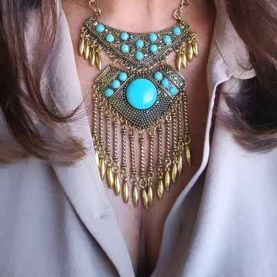 Colar dourado com pedras turquesa e pendentes metálicos sobre camisa bege