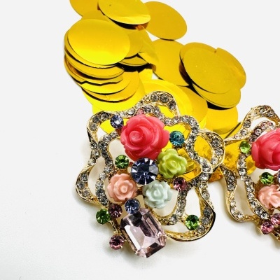 Brincos dourados com pedras e flores coloridas, fundo branco, confetes dourados