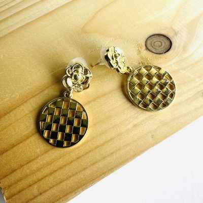 Brincos dourados com padrão quadriculado pendurados em base floral sobre madeira clara