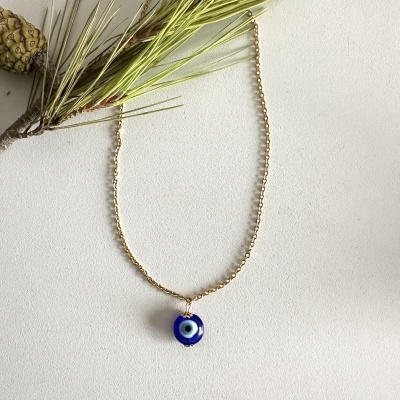 Colar com pendente azul em forma de olho com corrente dourada ao lado de ramo seco verde.