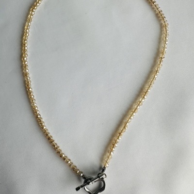 Colar com contas douradas e fecho em coração prateado sobre tecido branco.