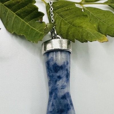 Colar com pendente de pedra azul e prata em forma de pé estilizado sobre fundo branco e folhas verdes