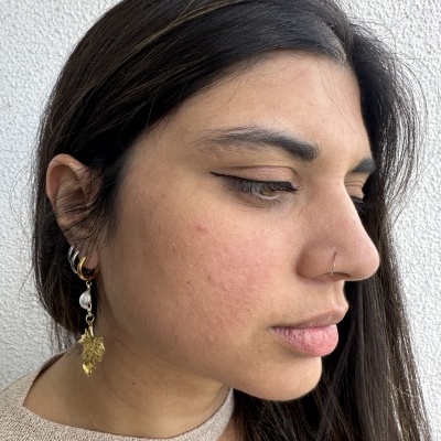 Mulher com brincos dourados com pérolas e pendente, vestido bege claro, pele morena e cabelo castanho.