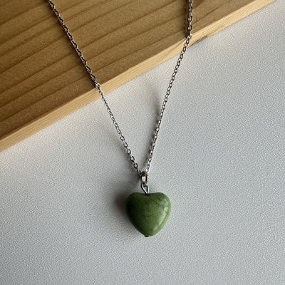 Colar com pendente em forma de coração verde e corrente metálica prateada sobre fundo branco e madeira clara