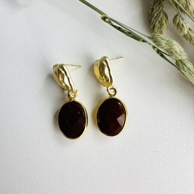 Brincos dourados com pedras bordô e ramos secos ao fundo