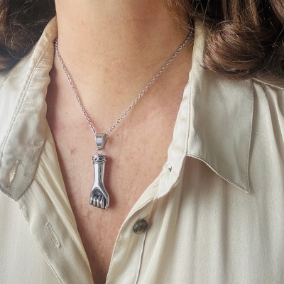 Colar prateado com pendente de mão em pescoço de pessoa com camisa bege clara