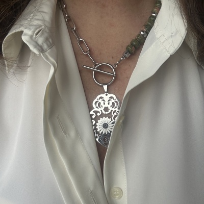 Colar com pendente metálico em padrão floral e corrente mista em elos metálicos e contas verdes sobre camisa branca.