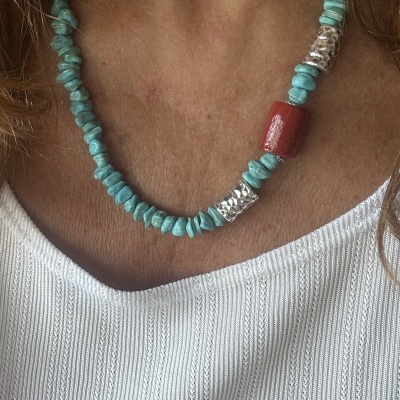 Colar de pedras turquesa, prateadas e vermelha num pescoço com camisa branca.