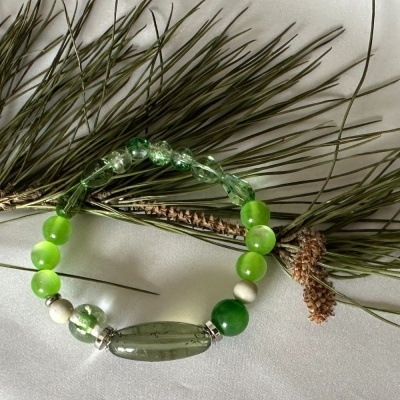 Pulseira verde e branca com várias contas dispostas sobre um tecido branco e raminhos de pinheiro