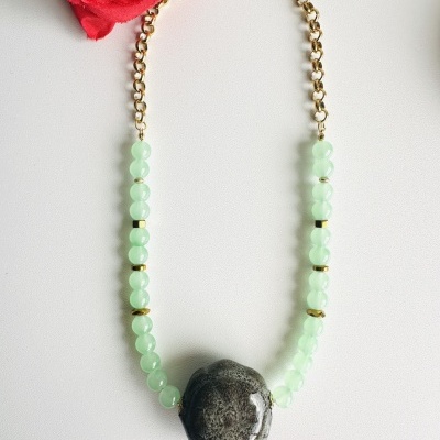 Colar com esferas verdes e pedra cinza escura sobre fundo branco com rosa vermelha.