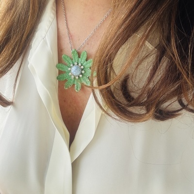 Colar com pendente em forma de flor verde com pedra azul claro usado por mulher com blusa branca