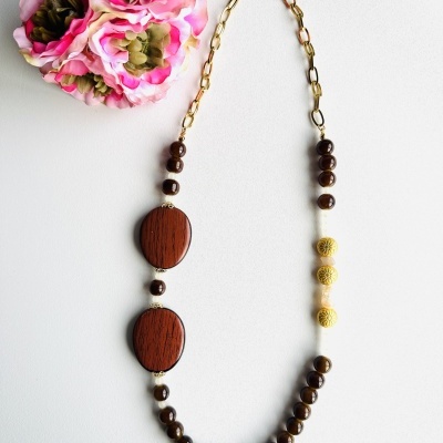 Colar com contas castanhas e douradas e peças ovais de madeira, em fundo branco com flores rosas e verdes.