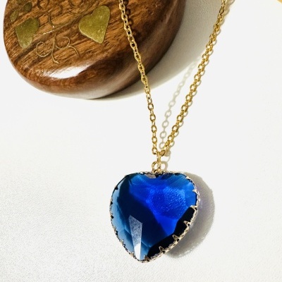 Colar com pendente azul coração e corrente dourada sobre superfície branca com caixa de madeira ao fundo