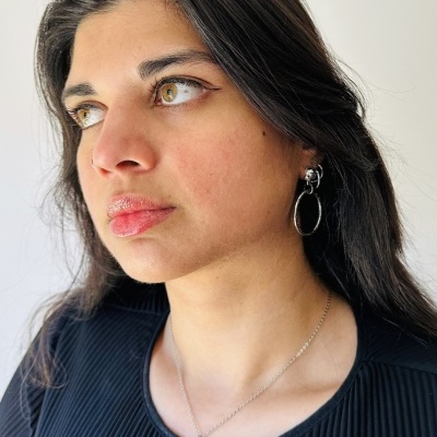 Mulher com colar prateado com pendente azul e cruz preta, brincos prateados de argolas e camisa preta texturizada