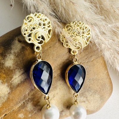 Brincos dourados com pedra azul e pérola pendurada sobre pedra e folhas secas