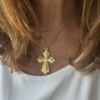Colar com pendente em forma de cruz dourada detalhada em pescoço de pessoa com blusa branca.