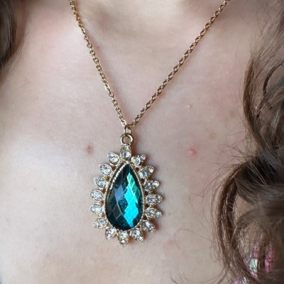 Colar dourado com pendente azul facetado e pedras pequenas ao redor no pescoço.