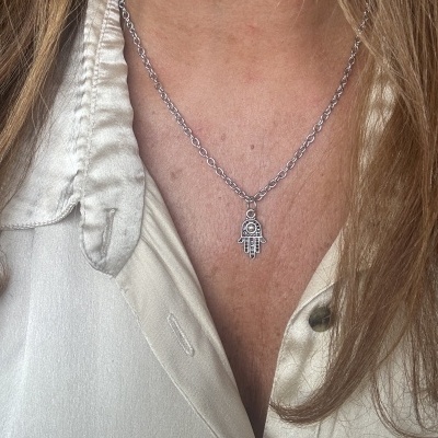 Colar com pendente em forma de mão prateada no pescoço de pessoa com camisa creme acetinada