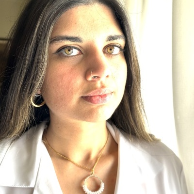 Mulher com colar dourado com pendente circular de pérolas e camisa branca perto de cortina clara