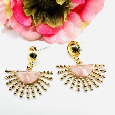Brincos dourados com pedra rosa e detalhes em pérolas formando raios perto de flor rosa