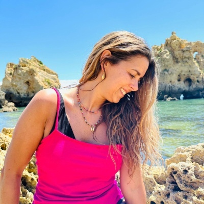 Mulher de top rosa e preto com colares coloridos e brincos dourados junto ao mar e rochas.