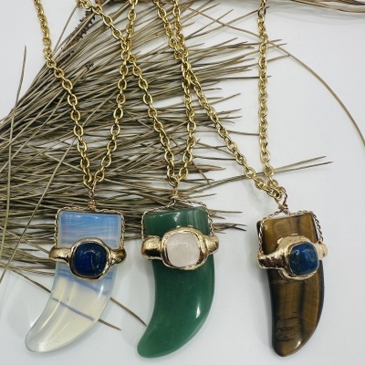 Três colares com pendentes em forma de dente e pedras coloridas sobre fundo branco e ramos secos