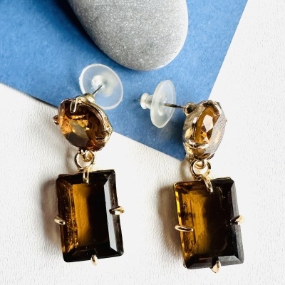 Brincos dourados com pedras castanhas em formato oval e retângulo, sobre fundo branco e azul com pedras cinzentas lisas.