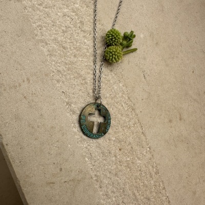 Colar de metal com pendente circular verde e cruz no centro sobre superfície bege